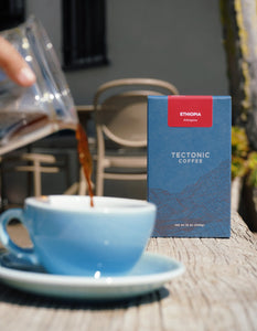 Blue cup of coffee with a Tectonic Coffee Ethiopia box on a wooden table.